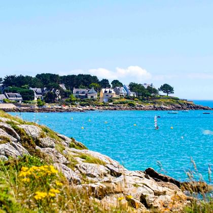 Felsige Küste mit gelben Wildblumen bei Saint-Colomban. Blaues Meer mit Booten und Bojen, weiße Häuser und Bäume am gegenüberliegenden Ufer.