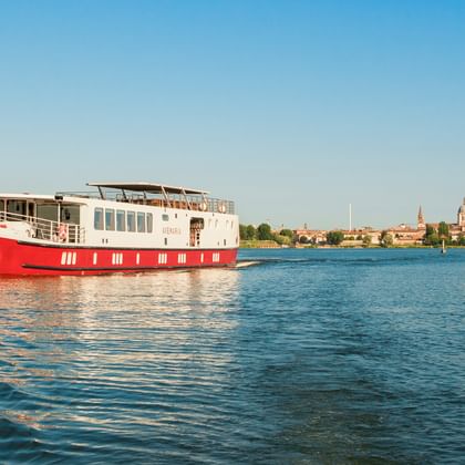 Rot-weißes Flusskreuzfahrtschiff MS Ave Maria auf ruhigem blauen Wasser mit Mantuas historischer Skyline im Hintergrund unter klarem Himmel.
