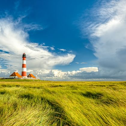 Leuchtturm Westerhever mit rot-weißen Streifen und angrenzenden Gebäuden in grünen Salzwiesen unter dramatischem blauen Himmel mit weißen Wolken.