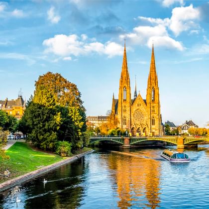 Gotische Kirche mit zwei Türmen in Straßburg neben einem Kanal mit Boot. Grüner Park und Brücke im Vordergrund unter blauem Himmel mit Wolken.