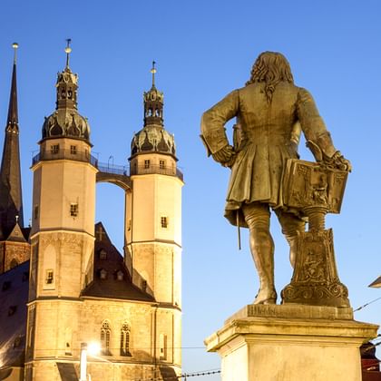 Bronzestatue eines Mannes mit Schwert auf dem Marktplatz in Halle an der Saale, mit beleuchteten Kirchtürmen vor blauem Abendhimmel.