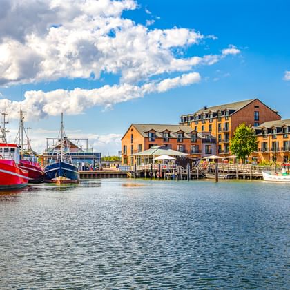 Heiligenhafener Hafen mit roten und blauen Fischerbooten am Kai. Bunte Gebäude säumen die Uferpromenade unter blauem Himmel mit Wolken.