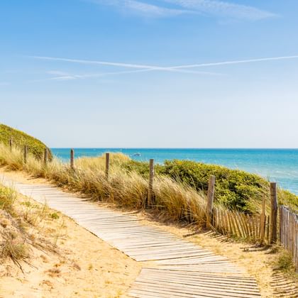 Holzsteg schlängelt sich durch Sanddünen mit Strandgras an der Pointe du Payre bei Jard-sur-Mer. Holzzaun säumt den Weg, türkisfarbenes Meer sichtbar.