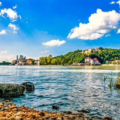 Blick über die Donau in Passau mit Kieselstrand, historischen Gebäuden und Dom links sowie einer Festung auf dem Hügel unter blauem Himmel.