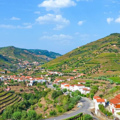 Dorf mit weißen Häusern und roten Dächern inmitten der terrassierten Weinberge des Douro-Tals. Eine kurvenreiche Straße führt durch grüne Hügel.