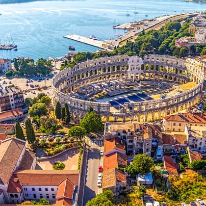 Luftaufnahme des antiken römischen Amphitheaters von Pula, umgeben von bunten Gebäuden mit roten Dächern. Der blaue Hafen mit Booten ist im Hintergrund sichtbar.