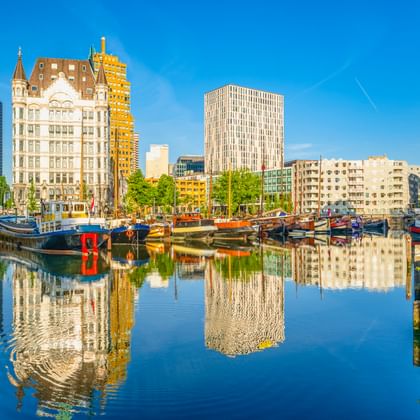 Rotterdamer Hafen mit Booten im ruhigen Wasser, das moderne Wolkenkratzer und das historische Witte Huis unter blauem Himmel spiegelt.
