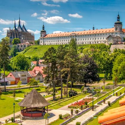 Panoramablick auf Kutná Hora mit gotischem Dom, barockem Schloss, terrassierten Gärten und Park mit Holzpavillon unter blauem Himmel.