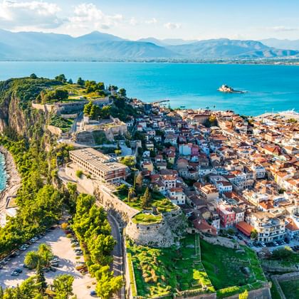 Luftaufnahme der Halbinsel Nafplio mit historischer Festung, rotgedeckten Gebäuden und türkisfarbener Bucht. Berge im Hintergrund unter blauem Himmel.