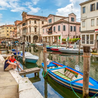Radfahrer mit roten Tourenrädern rasten an einem Kanal in einer italienischen Stadt. Bunte Boote liegen an Holzpfählen vertäut, historische Gebäude säumen den Kanal.