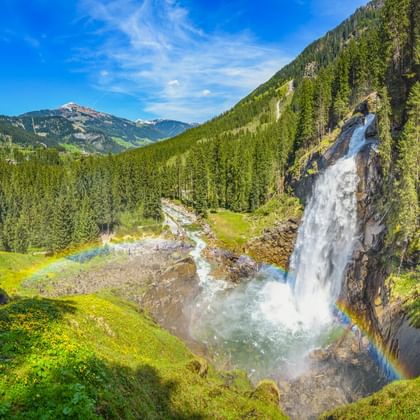Krimmler Wasserfälle stürzen über Felsklippe mit Regenbogen im Sprühnebel, umgeben von grünen Almwiesen und dichtem Wald in Österreich.