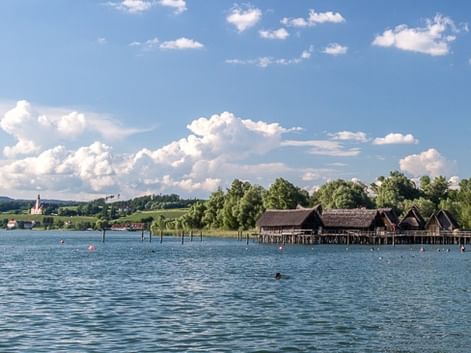 Rekonstruierte prähistorische Pfahlbauten am Bodensee mit Kirchturm in der Ferne. Blaues Wasser im Vordergrund, grüne Landschaft und Himmel.