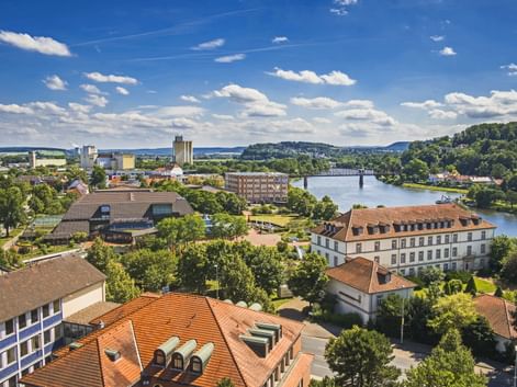 Luftaufnahme von Hameln mit rotgedeckten Gebäuden, der Weser, einer Brücke und grünen Hügeln unter blauem Himmel mit Wolken.