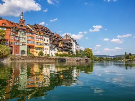 Historische bunte Fachwerkhäuser der Laufenburger Altstadt spiegeln sich im ruhigen Flusswasser. Kirchturm ragt über den Gebäuden auf.