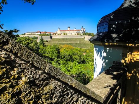 Blick auf die Festung Marienberg in Würzburg von Steinbefestigungen, mit Weinbergen am Hang und blauem Himmel. Historischer Turm im Vordergrund.