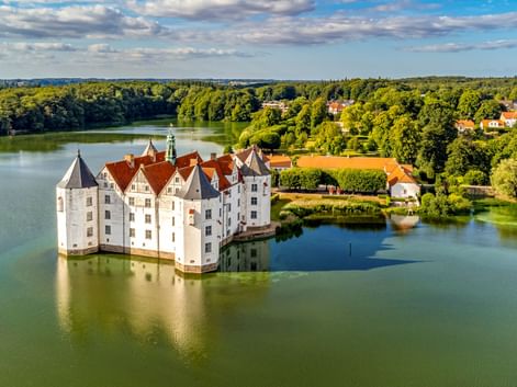 Schloss Glücksburg, ein weißes Renaissance-Wasserschloss mit roten Dächern, umgeben von einem See. Grüne Wälder und blauer Himmel im Hintergrund.