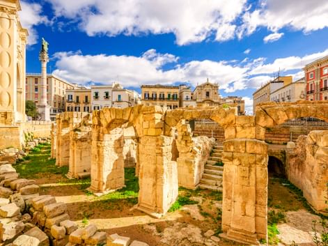 Ruinen des antiken römischen Amphitheaters in Lecce mit Steinsäulen und Bögen, umgeben von historischen Gebäuden unter blauem Himmel.