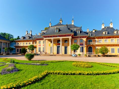 Schloss Pillnitz mit gelber Fassade und weißen Säulen, umgeben von gepflegten Rasenflächen mit bunten Blumenbeeten unter blauem Himmel.