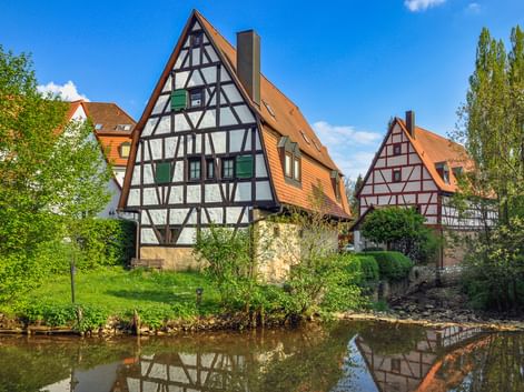 Traditionelle bayerische Fachwerkhäuser mit orangen Ziegeldächern spiegeln sich in einem ruhigen Fluss in Hersbruck, umgeben von grünen Bäumen.