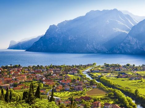 Dorf Pietramurata im Sarcatal mit rotgedeckten Häusern, grünen Feldern und Gardasee, umgeben von dramatischen Berggipfeln unter blauem Himmel.