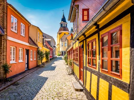 Kopfsteinpflasterstraße in Faaborgs Altstadt mit bunten Fachwerkhäusern in Gelb, Orange und Rot. Ein Kirchenglockenturm erhebt sich im Hintergrund.
