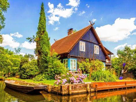 Blaues Holzhaus mit orangefarbenem Ziegeldach am Wasser in Lübben Spreewald. Holzsteg mit Blumen und Boot davor, umgeben von Bäumen.