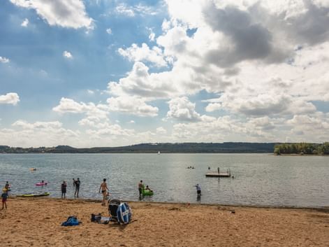 Sandstrand am Brombachsee mit schwimmenden und spielenden Menschen im Wasser. Blauer Himmel mit weißen Wolken über dem See und bewaldeter Uferlinie.