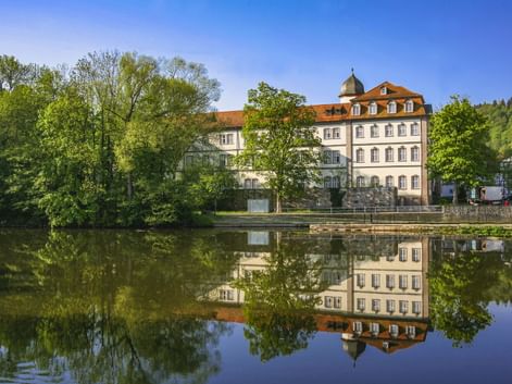 Weißes Barockschloss in Rotenburg an der Fulda mit orangefarbenem Dach und Turm, spiegelt sich im ruhigen Flusswasser. Grüne Bäume rahmen das Gebäude unter blauem Himmel.
