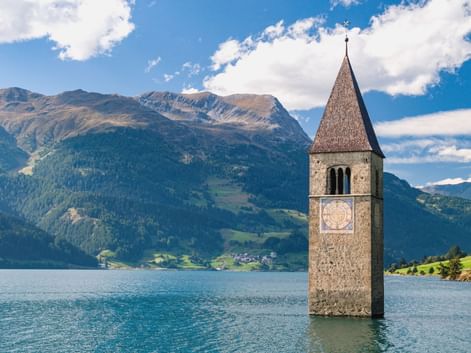 Historischer Kirchturm von Graun ragt aus dem Reschensee in Bayern, mit Bergen und blauem Himmel im Hintergrund entlang der Via Claudia Augusta.