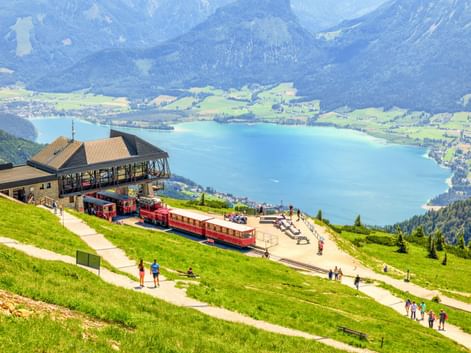 Rote Zahnradbahnen an der Schafberg Bergstation auf grüner Almwiese mit Blick auf den türkisen Wolfgangsee und Berge in Österreich.
