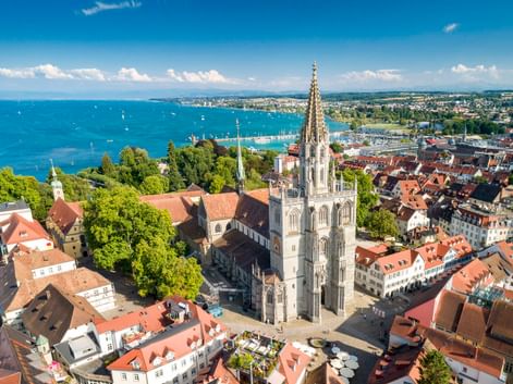 Luftaufnahme des Konstanzer Münsters mit gotischem Turm, umgeben von Gebäuden mit roten Dächern und dem Bodensee im Hintergrund.
