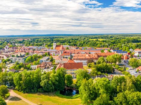 Luftaufnahme von Lübben mit roten Ziegeldächern, weißem Kirchturm, grünen Parks und Wäldern unter teilweise bewölktem Himmel.