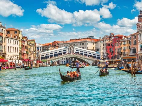 Die berühmte Rialtobrücke überspannt den Canal Grande in Venedig mit Gondeln und Booten im Wasser. Bunte historische Gebäude säumen beide Seiten unter blauem Himmel.