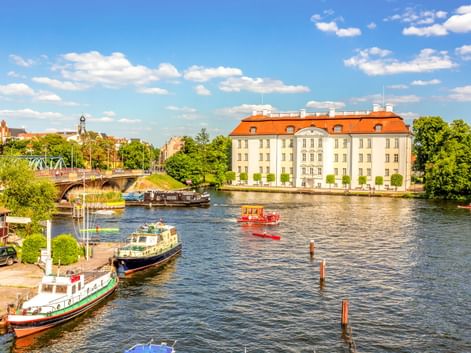 Schloss Köpenick mit orangefarbenem Dach neben einem Fluss in Berlin. Boote am Ufer vertäut, Brücke und Kirchtürme im Hintergrund sichtbar.