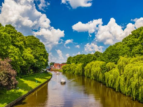 Fluss Ems in Meppen fließt zwischen üppig grünen Bäumen und Weiden. Zwei Boote fahren auf dem ruhigen Wasser unter blauem Himmel mit weißen Wolken.