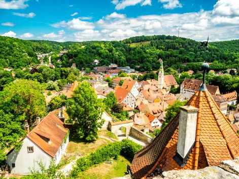 Luftaufnahme von Pappenheim mit roten Ziegeldächern, Kirchturm und umliegenden grünen Hügeln unter blauem Himmel mit weißen Wolken.