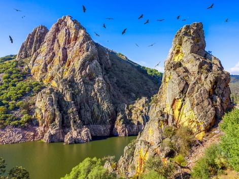 Dramatische Felsformationen am Castillo de Monfragüe, die aus einem Stausee aufragen. Vögel kreisen über den Klippen unter blauem Himmel.