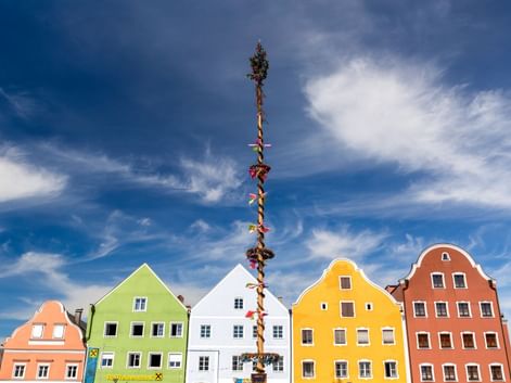 Hoher geschmückter Maibaum vor bunten Giebelhäusern in Obernberg am Inn. Gebäude in Grün, Weiß, Gelb und Rot unter blauem Himmel.