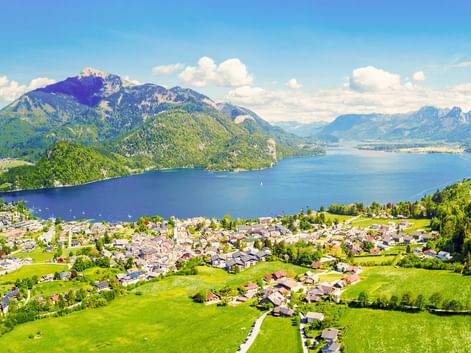 Luftaufnahme von St. Gilgen mit grünen Wiesen am Wolfgangsee, umgeben von bewaldeten Bergen unter blauem Himmel in Österreich.