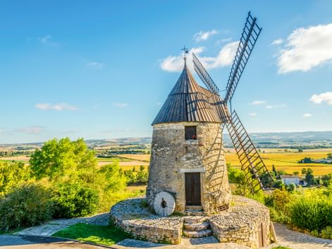 Steinwindmühle mit blauem Kegeldach und Holzflügeln in Castelnaudary, mit Blick auf hügelige Landschaft mit Feldern unter blauem Himmel.