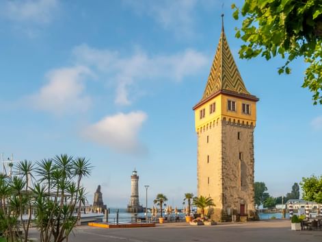 Historischer Mangturm mit buntem Ziegeldach am Lindauer Hafeneingang am Bodensee. Leuchtturm im Hintergrund, Palmen im Vordergrund.