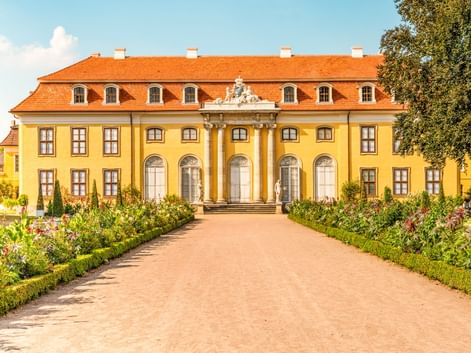 Gelbes Barockschloss mit orangefarbenem Dach und weißen Säulen in Mosigkau bei Dessau. Ein Kiesweg führt durch einen formalen Garten zum Eingang.