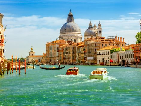 Boote auf dem Canal Grande in Venedig mit den Kuppeln der Basilika Santa Maria della Salute im Hintergrund. Historische Gebäude säumen den Kanal.