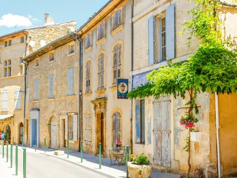 Sonnige Straße in Aix-en-Provence mit traditionellen Steingebäuden, pastellfarbenen Fensterläden und grünen Bäumen. Grüne Poller säumen den Fußgängerbereich.