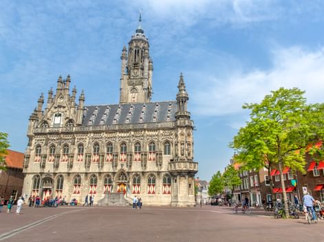 Gotisches Rathaus von Middelburg mit verzierter Fassade und hohem Turm auf einem Platz. Menschen und Radfahrer davor, flankiert von grünen Bäumen.