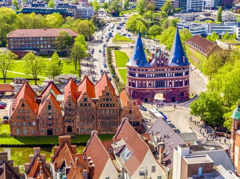 Luftaufnahme des Lübecker Holstentors mit Zwillingstürmen und roten Backsteingebäuden. Grüne Parks und Stadtstraßen umgeben das Wahrzeichen.