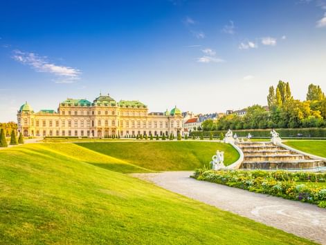 Schloss Belvedere in Wien mit gepflegten Barockgärten, weißen Statuen, Brunnen und grünen Rasenflächen unter blauem Himmel.