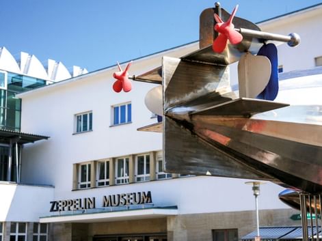Weißes modernistisches Gebäude mit Zeppelin Museum Eingangsschild und großer Metall-Propeller-Skulptur mit roten Akzenten über dem Eingang.