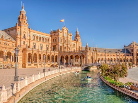 Plaza de España in Sevilla zeigt verzierte Backsteingebäude mit Türmen und Bögen entlang eines Kanals mit Brücke. Besucher flanieren über den Platz.