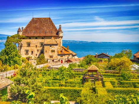 Mittelalterliches Schloss in Yvoire mit Steinmauern und rotem Ziegeldach mit Blick auf den Genfersee. Formaler Garten mit geometrischen Hecken im Vordergrund.